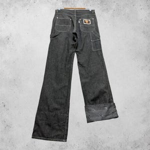 Könnte beinhalten: Dunkelgraue Jeans aus Denim mit weitem Bein. Die Jeans haben einen klassischen Fünf-Taschen-Stil, einen kleinen Logoaufnäher auf der Rückseite und dekorative Nähte. Die Manschetten sind umgeschlagen und zeigen ein kontrastierendes Detail.