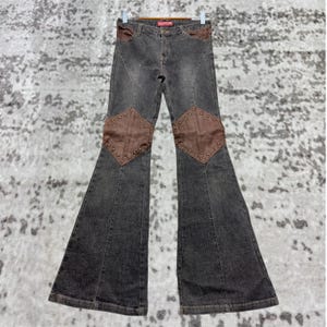 Könnte beinhalten: Dunkelgraue Schlaghose mit braunen Wildlederaufnähern an den Knien und oberen Gesäßtaschen. Die Aufnäher sind mit kleinen, silberfarbenen Nieten besetzt. Die Jeans hängt an einem Kleiderbügel.