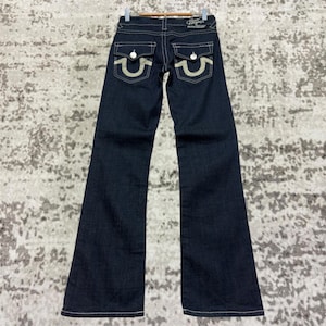 Könnte beinhalten: Dunkelblaue Jeans mit ausgestelltem Bein. Die Jeans hat hellfarbene Nähte, Gesäßtaschen mit geschwungenem Design und ein Markenetikett am Bund. Die Jeans wird auf einem Kleiderbügel präsentiert.