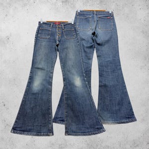 Könnte beinhalten: Zwei Paar ausgestellte Jeans aus blauem Denim. Das vordere Paar hat einen Knopfverschluss, zwei aufgesetzte Vordertaschen und eine verwaschene Optik. Das hintere Paar zeigt die Rückansicht mit zwei aufgesetzten Taschen und einem Bund.