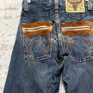 May include: Close-up of blue denim jeans with brown suede pockets and zippers. The back pockets have decorative stitching. A leather patch with the text "EDWIN" and a bull graphic is visible.