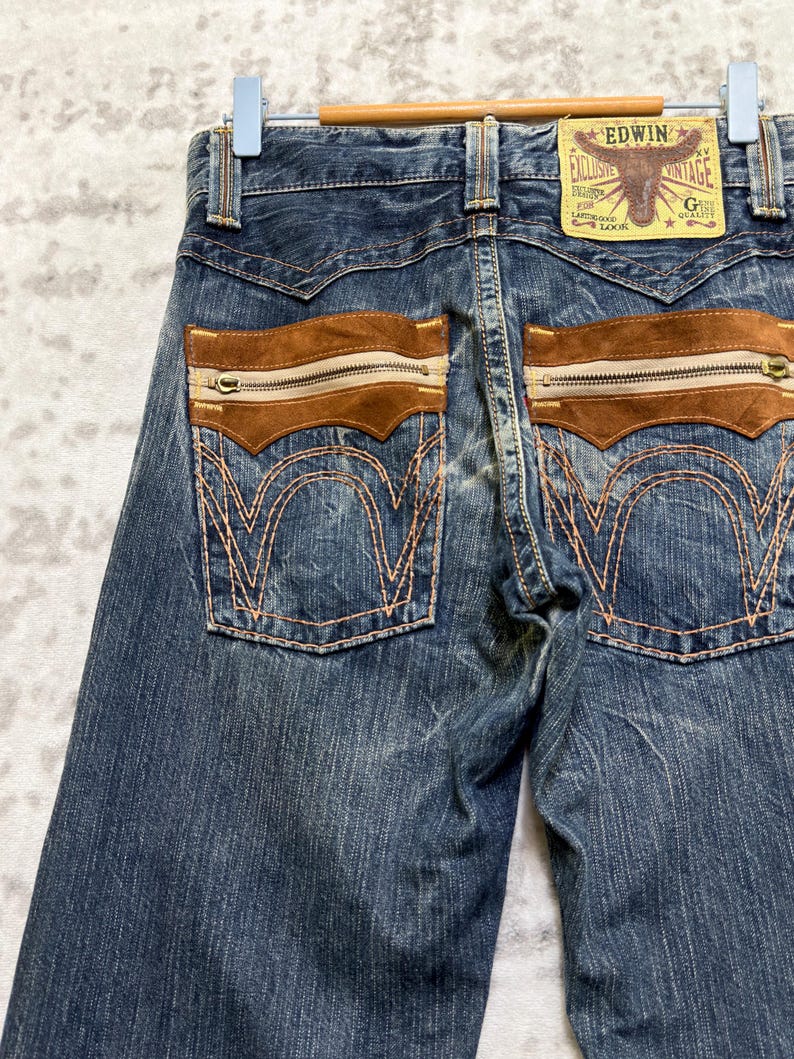 May include: Close-up of blue denim jeans with brown suede pockets and zippers. The back pockets have decorative stitching. A leather patch with the text "EDWIN" and a bull graphic is visible.