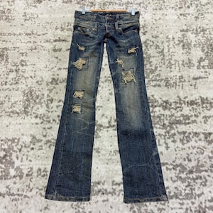 Könnte beinhalten: Verwaschene blaue Jeans mit mehreren zerrissenen Patches. Die Jeans hat eine niedrige Taille, einen Reißverschluss und ein ausgestelltes Bein. Die Jeans hängt an einem Holzbügel.