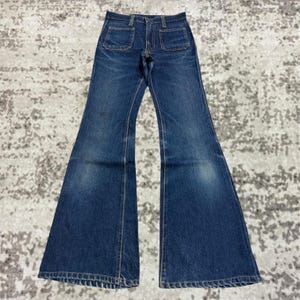 Könnte beinhalten: Dunkelblaue Schlaghose aus Denim mit zwei Vordertaschen. Die Jeans hat eine hohe Taille und einen ausgestellten Bein-Design. Der Denim hat ein verwaschenes Aussehen, mit helleren Bereichen an den Oberschenkeln und Knien. Die Jeans liegt flach auf einer gemusterten Oberfläche.