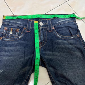 May include: Dark blue denim jeans with a green measuring tape showing the waist size. The jeans have a button closure, front pockets, and decorative stitching. The waist measures approximately 13 inches (33 cm).