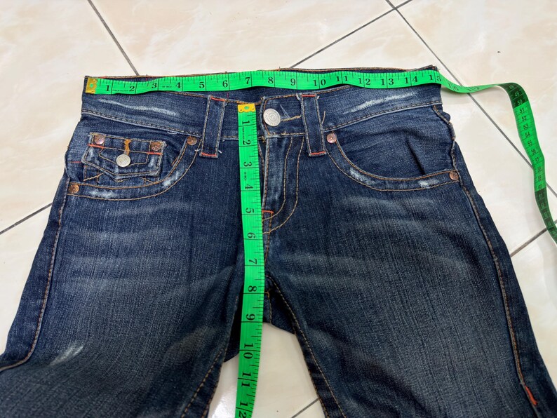 May include: Dark blue denim jeans with a green measuring tape showing the waist size. The jeans have a button closure, front pockets, and decorative stitching. The waist measures approximately 13 inches (33 cm).