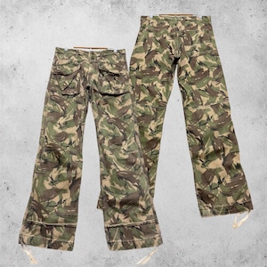 Könnte beinhalten: Zwei Hosen mit Camouflage-Muster in Grün-, Braun- und Beigetönen. Die Hosen verfügen über Vordertaschen mit Klappen, einen geknöpften Hosenschlitz und verstellbare Kordelzüge an den unteren Säumen der Hosenbeine.