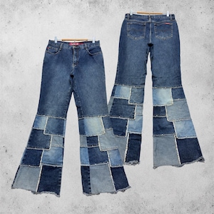 May include: Blue denim jeans with a patchwork design on the flared legs. The jeans are displayed on a hanger, showcasing the front and back views. The patchwork consists of various shades of blue denim.