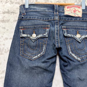 May include: Dark wash denim jeans with back pockets featuring decorative stitching and silver button closures. The jeans have a distressed look with a True Religion label on the back. The jeans are hanging on a blue hanger.