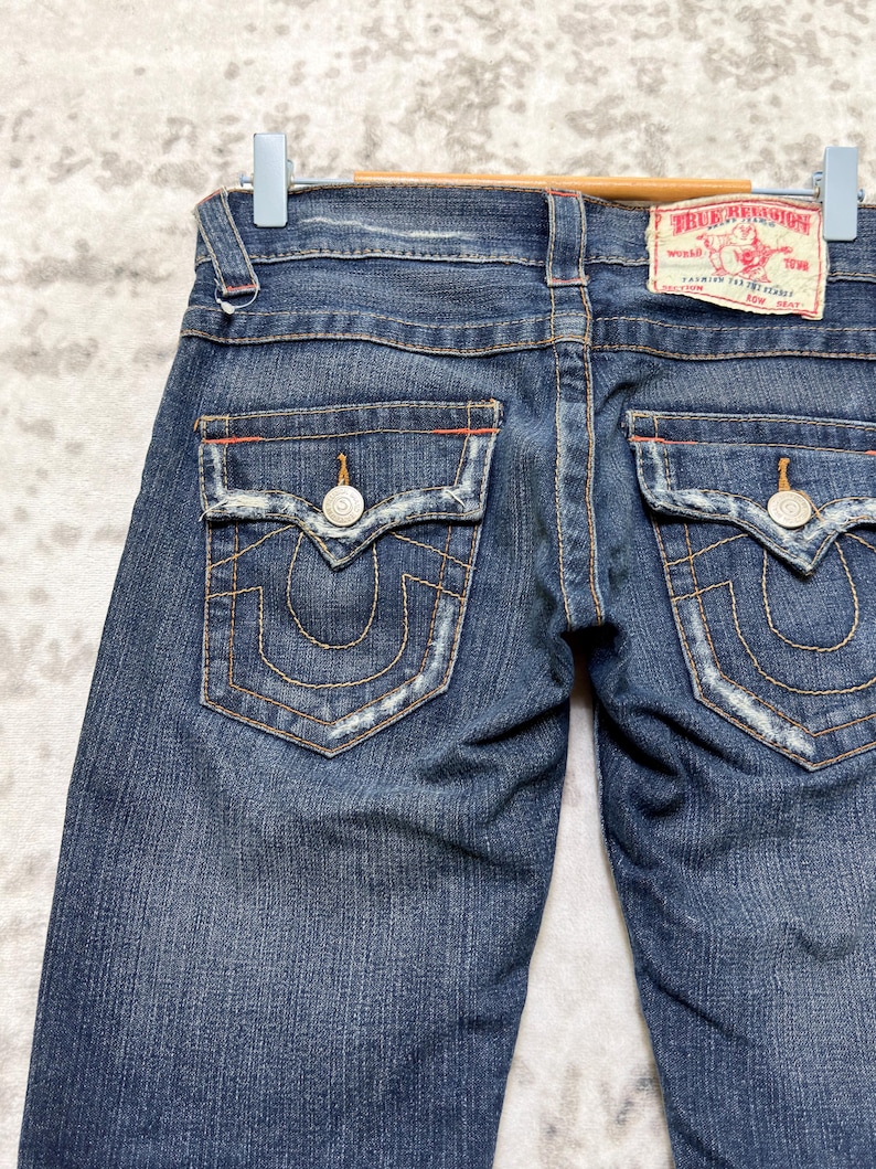 May include: Dark wash denim jeans with back pockets featuring decorative stitching and silver button closures. The jeans have a distressed look with a True Religion label on the back. The jeans are hanging on a blue hanger.