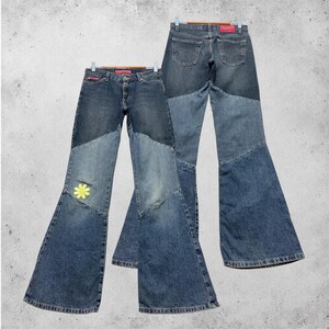 Könnte beinhalten: Zwei Jeanshosen mit Patchwork-Denim-Schlaghose. Die Jeans haben eine Mischung aus hell- und dunkelblauen Denim-Einsätzen. Ein Paar hat eine gelbe Gänseblümchen-Applikation auf der Vorderseite. Die Jeans hängen an Holzbügeln.