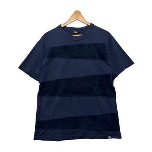 Könnte beinhalten: Marineblaues Kurzarm-T-Shirt mit horizontalen Streifen aus dunklerem, marineblauem, samtigem Material. Das Shirt hängt an einem Holzbügel. Das Shirt hat einen Rundhalsausschnitt und ein kleines Etikett unten rechts.