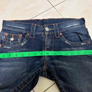 May include: Dark blue denim jeans with a button closure and multiple pockets. The jeans are being measured with a green measuring tape. The waist measures approximately 16 inches across.