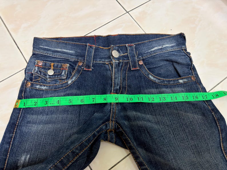 May include: Dark blue denim jeans with a button closure and multiple pockets. The jeans are being measured with a green measuring tape. The waist measures approximately 16 inches across.