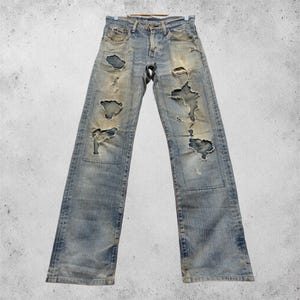 Puede incluir: Vaqueros de mezclilla azul claro con detalles desgastados, con grandes rasgaduras y parches. Los vaqueros est&aacute;n colgados de una percha, mostrando una est&eacute;tica vintage y desgastada. El lavado descolorido y el dise&ntilde;o rasgado dan a los vaqueros un aspecto &uacute;nico y atrevido.