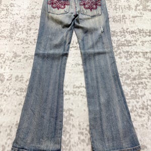 May include: Light blue denim jeans with a flared leg design. The back pockets feature a decorative embroidered floral pattern in shades of red. The jeans are displayed on a wooden hanger.