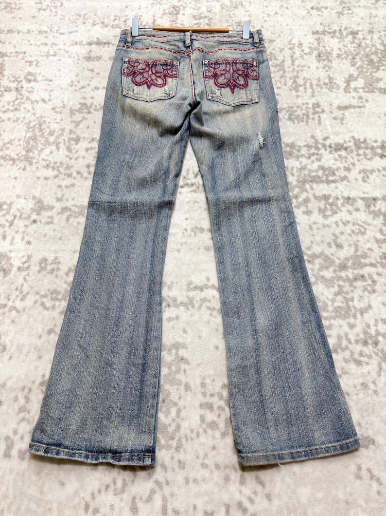 May include: Light blue denim jeans with a flared leg design. The back pockets feature a decorative embroidered floral pattern in shades of red. The jeans are displayed on a wooden hanger.