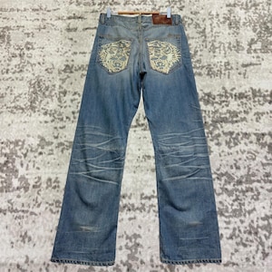 Può includere: Jeans di denim blu chiaro con disegni di tigre ricamati sulle tasche posteriori. I jeans hanno un classico design a cinque tasche e una vestibilità dritta. I jeans sono appesi a una gruccia di legno.
