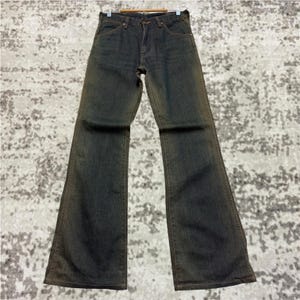 May include: Dark wash denim jeans with a flared leg. The jeans have a classic five-pocket design and a button and zipper closure. The denim has a faded, worn look with subtle brown accents along the seams and edges, giving them a vintage aesthetic.