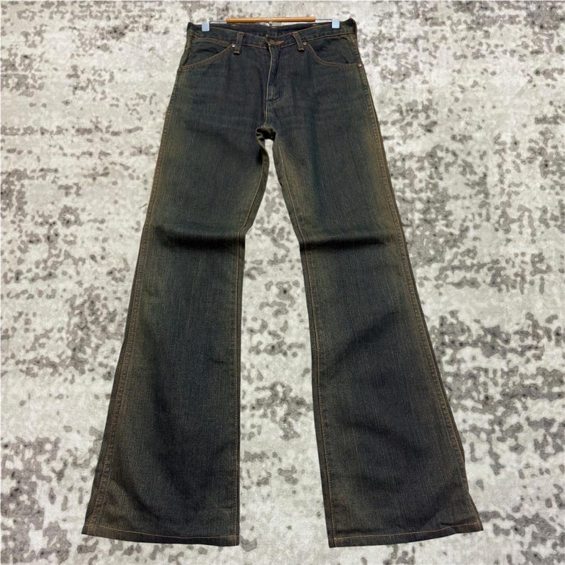 May include: Dark wash denim jeans with a flared leg. The jeans have a classic five-pocket design and a button and zipper closure. The denim has a faded, worn look with subtle brown accents along the seams and edges, giving them a vintage aesthetic.