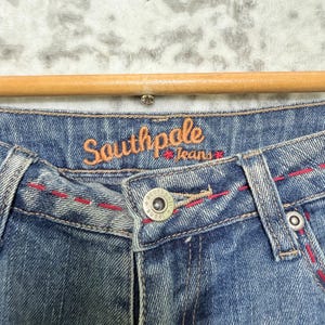 May include: Close-up of blue denim jeans with red stitching details. The jeans feature a button closure and the brand name "Southpole Jeans" in orange script.