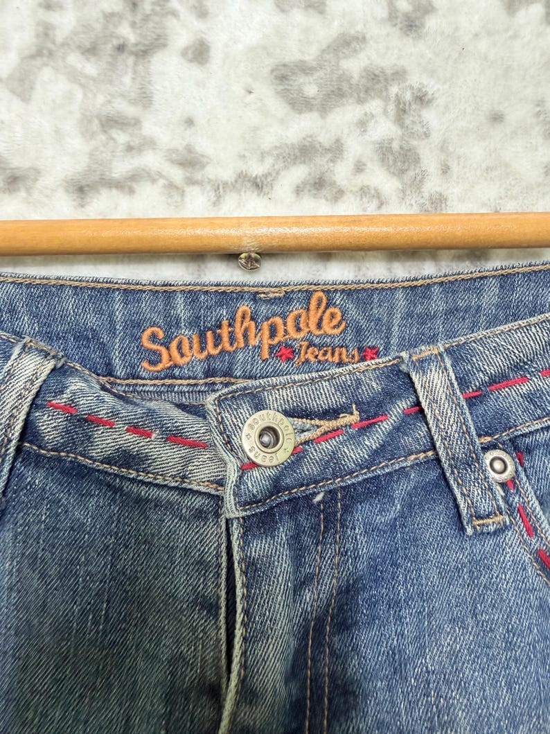 May include: Close-up of blue denim jeans with red stitching details. The jeans feature a button closure and the brand name "Southpole Jeans" in orange script.