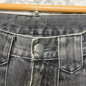 May include: Close-up of the waistband of black denim jeans. The jeans have a silver button with the brand name "Wrangler" and a silver rivet. The jeans are hanging on a wooden hanger.