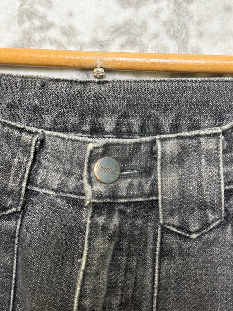May include: Close-up of the waistband of black denim jeans. The jeans have a silver button with the brand name "Wrangler" and a silver rivet. The jeans are hanging on a wooden hanger.