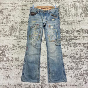 Könnte beinhalten: Hellblaue Jeans mit dekorativen Patches und farbigen Nähten. Die Jeans haben einen Bootcut-Stil und tragen den Text "Back Number" auf dem Bund. Auf der Vorderseite der Jeans sind Patches aufgenäht.