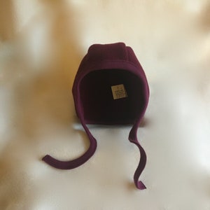 May include: A burgundy fleece baby bonnet with a tie at the back. The bonnet has a small white tag with the text "Made with love" on it.