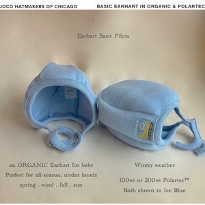 May include: Two baby hats, one made of organic cotton and one made of Polartec fleece, both in a light blue color. The text "Earhart Basic Pilots" is visible above the hats. The text "an ORGANIC Earhart for baby" is below the cotton hat. The text "Wintry weather" is below the fleece hat. The text "100wt or 200wt Polartec™" is below the fleece hat. The text "Both shown in Ice Blue" is below the fleece hat.