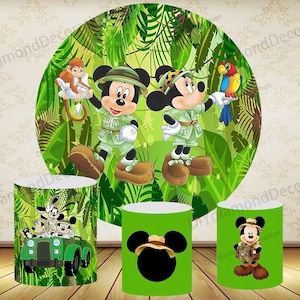 May include: A jungle-themed party decoration set featuring Mickey Mouse and friends. The set includes a large circular backdrop with Mickey and a monkey, and three cylindrical stands with various character designs. The color scheme is predominantly green and brown.