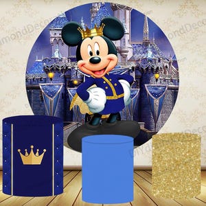 May include: A party backdrop featuring Mickey Mouse in a royal outfit, with a castle background. Three cylindrical pedestals are in the foreground: one blue, one gold glitter, and one navy with a gold crown design.