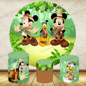 Könnte beinhalten: Ein farbenfrohes Party-Dekorationsset zum Thema Dschungel mit Micky Maus, Minnie Maus, Pluto, Goofy und Donald Duck in Safari-Outfits. Das Set enthält eine große kreisförmige Kulisse und drei zylindrische Podeste.