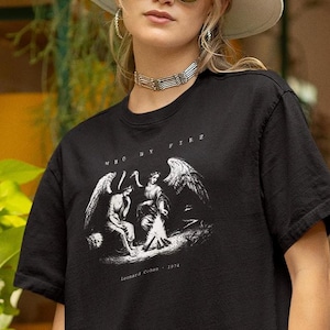 May include: Black t-shirt with a graphic of two figures with wings, and the text "WAY BY FIRE" and "Leonard Cohen - 1974". The person is wearing a white hat, sunglasses, and a silver choker.