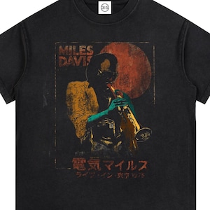 May include: A black short-sleeve t-shirt featuring a graphic print of Miles Davis playing a trumpet. The design includes a large red circle, the text "MILES DAVIS," and Japanese characters that read "Denki Miles" and "Live in Tokyo 1975." The shirt has a crew neckline.
