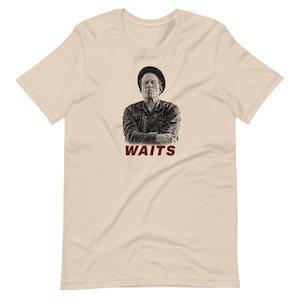 May include: A beige t-shirt with a black and white image of Tom Waits wearing a hat and a dark jacket. The text "WAITS" is printed in red below the image.