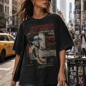 May include: A charcoal black oversized t-shirt featuring a graphic print with the text CONTEMPORARY ART WORLD NEW YORK CITY 1993, a portrait of a person with a blue stripe across the eyes, a red chair, a yellow banana, and the name Basquiat.