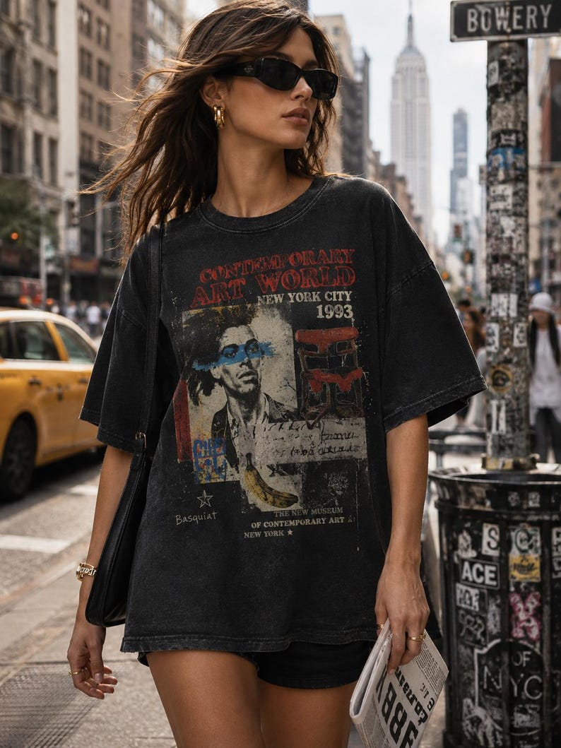May include: A charcoal black oversized t-shirt featuring a graphic print with the text CONTEMPORARY ART WORLD NEW YORK CITY 1993, a portrait of a person with a blue stripe across the eyes, a red chair, a yellow banana, and the name Basquiat.