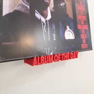 May include: A red vinyl record holder with the words "Album of the Day" printed on it. The holder is attached to a wall and a vinyl record is displayed on it. The record cover features a black and white photo of a man with the words "Public Enemy" printed on it.