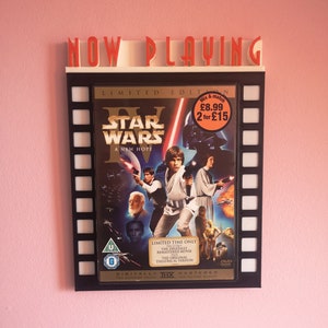 May include: A black framed movie poster with a film strip border. The poster features the Star Wars IV: A New Hope movie cover art. The poster is displayed on a pink wall with a sign that reads "NOW PLAYING".