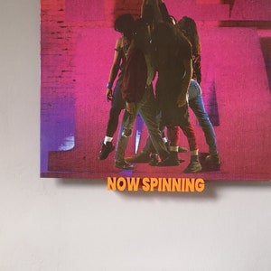 May include: A black vinyl record player with a black turntable and a black tonearm. The record player is playing a Pearl Jam record album. The album cover features a group of five people in a pink and purple background. The text "Pearl Jam" is printed in white on the album cover. The text "Now Spinning" is printed in orange below the album cover.