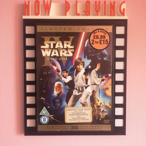 May include: A black and white movie poster for Star Wars: A New Hope. The poster features the main characters, including Luke Skywalker, Princess Leia, Han Solo, and Darth Vader. The poster is framed in a black frame with a film strip design. The text "NOW PLAYING" is displayed above the poster.