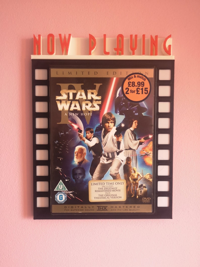 May include: A black and white movie poster for Star Wars: A New Hope. The poster features the main characters, including Luke Skywalker, Princess Leia, Han Solo, and Darth Vader. The poster is framed in a black frame with a film strip design. The text "NOW PLAYING" is displayed above the poster.
