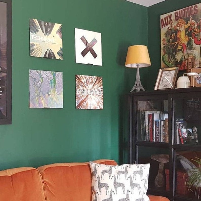 A square of four vinyl records is seen displayed against a green wall above a brown couch. A cabinet holds books and a lamp shaped like the Eifel Tower.