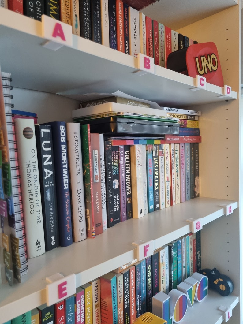 May include: A white bookshelf with books organized by color and labeled with letters. The top shelf has a red UNO card game box. The bottom shelf has a rainbow LOVE sign.