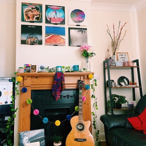 May include: A living room interior with a fireplace decorated with a garland of colorful pom poms. A wooden acoustic guitar leans against the fireplace. A black leather sofa with a red throw pillow is in the foreground. A shelf with various items is in the background. The wall above the fireplace is decorated with framed vinyl records, including albums by Grease, Harry Styles, and ABBA.