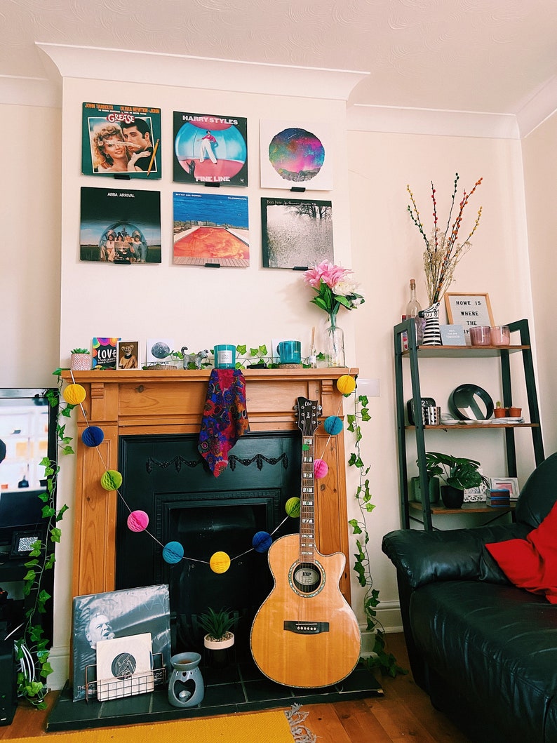 May include: A living room interior with a fireplace decorated with a garland of colorful pom poms. A wooden acoustic guitar leans against the fireplace. A black leather sofa with a red throw pillow is in the foreground. A shelf with various items is in the background. The wall above the fireplace is decorated with framed vinyl records, including albums by Grease, Harry Styles, and ABBA.