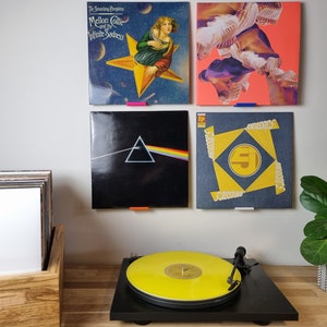 May include: Four vinyl records are displayed on a wall, with a turntable and a plant on a wooden surface below. The records are "The Smashing Pumpkins Mellon Collie and the Infinite Sadness", "Dark Side of the Moon", "EP", and an abstract design. The turntable has a yellow record on it.