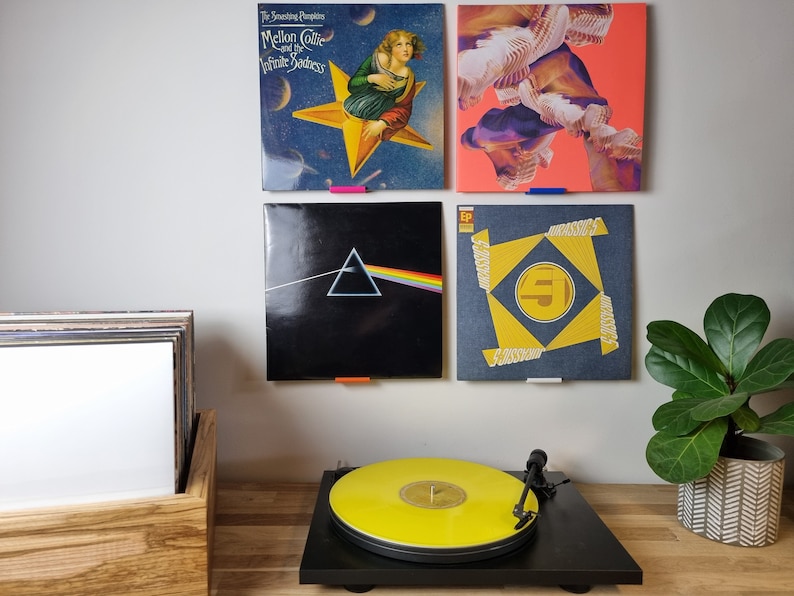 May include: Four vinyl records are displayed on a wall, with a turntable and a plant on a wooden surface below. The records are "The Smashing Pumpkins Mellon Collie and the Infinite Sadness", "Dark Side of the Moon", "EP", and an abstract design. The turntable has a yellow record on it.